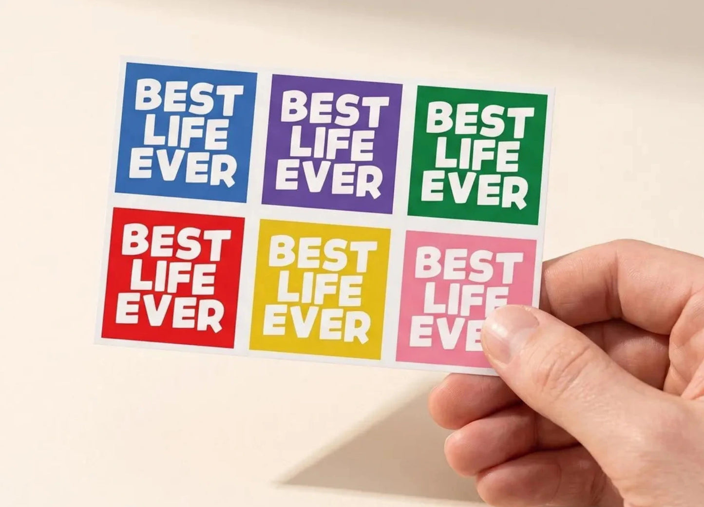 A hand holds a sheet of six square stickers with the phrase 'BEST LIFE EVER' in white text on colorful backgrounds. The stickers are arranged in a 2x3 grid with blue, purple, green, red, yellow, and pink colors.
