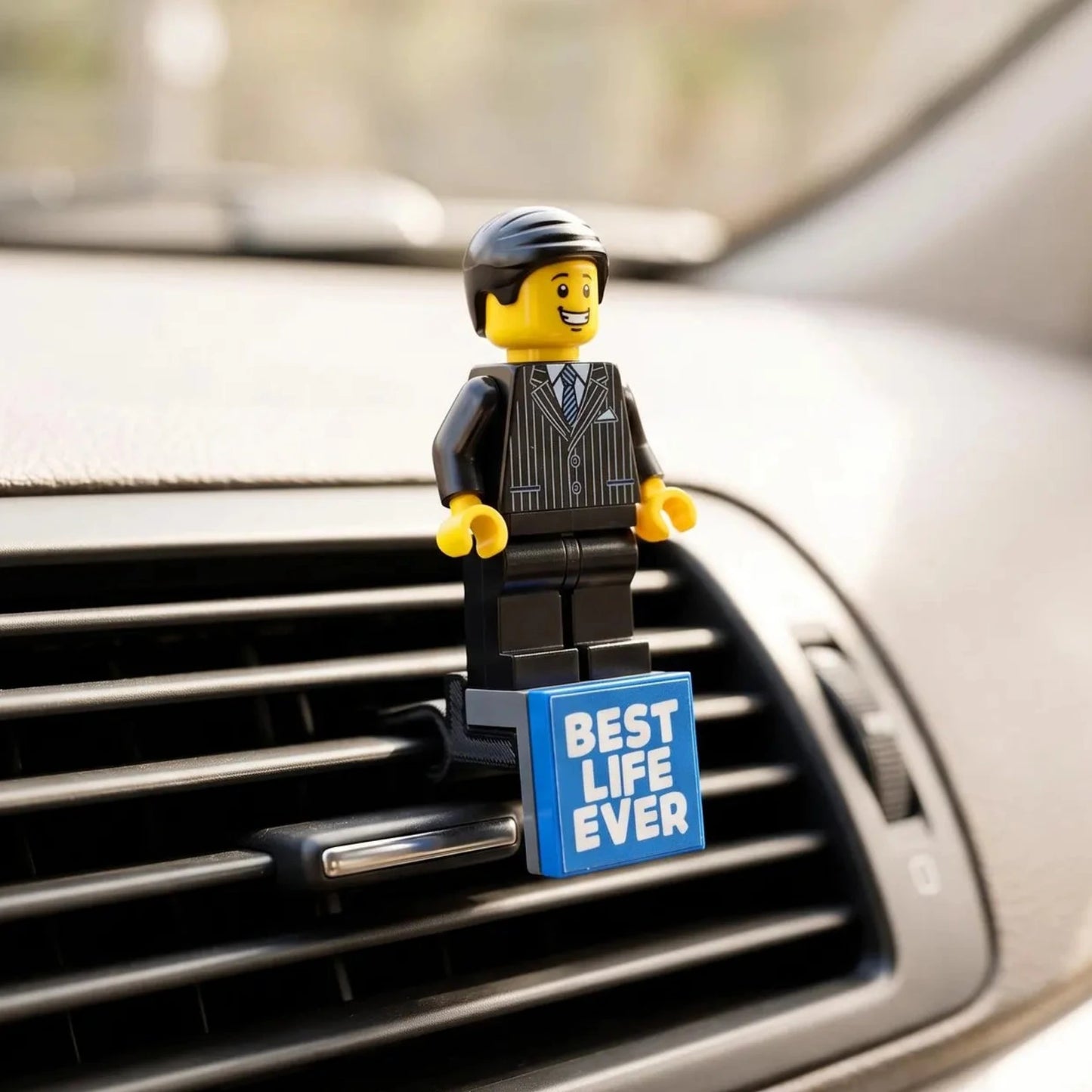 A small LEGO-style figurine in a black pinstripe suit stands on a blue sign that reads “BEST LIFE EVER,” placed on a car air vent.