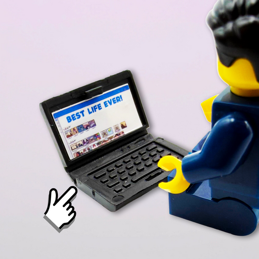 A small black toy laptop sits open on a pale gray background, with a hand pointing at the screen. A LEGO minifigure viewed from behind is seated to the right, looking at the laptop.