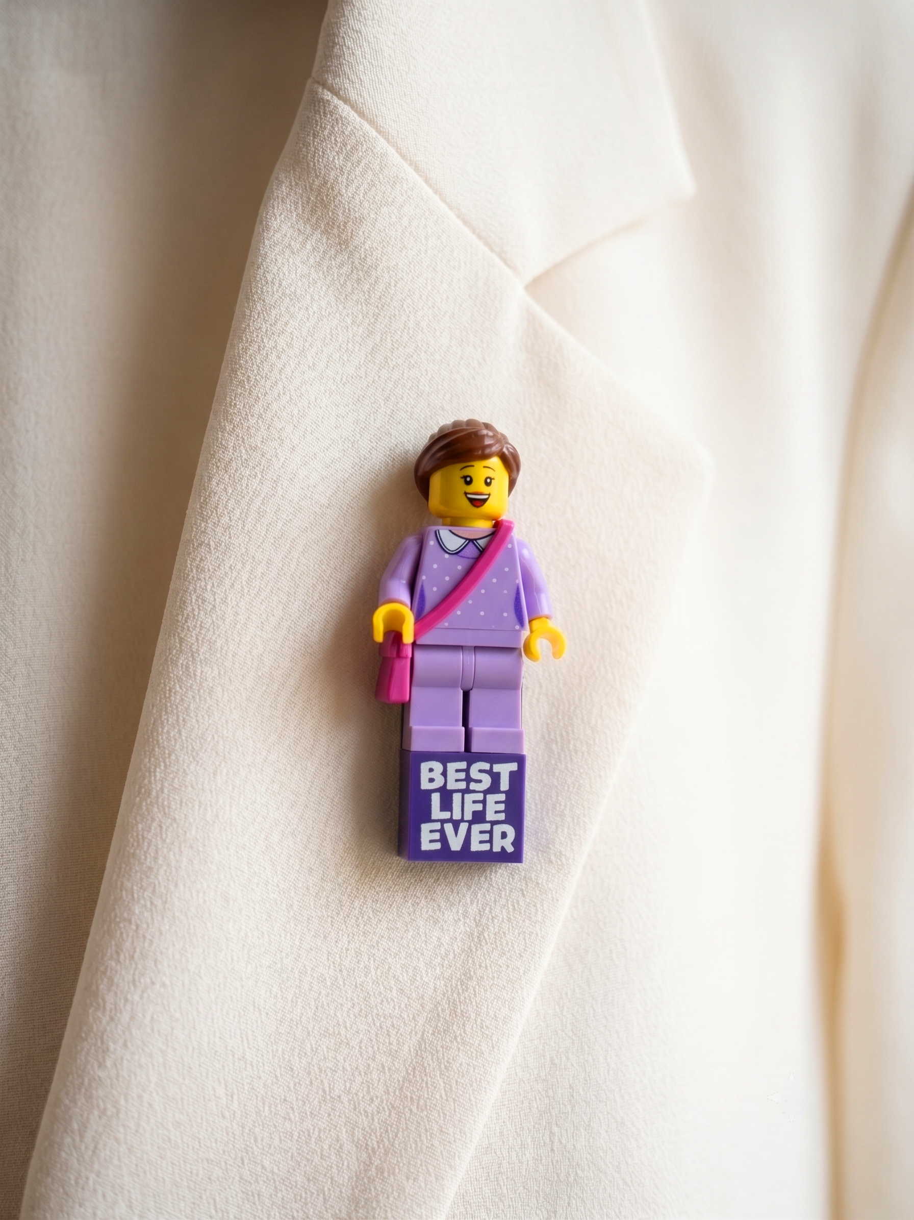 A small LEGO minifigure pin styled as a woman in a lavender suit with a pink purse, pinned to a cream-colored blazer. The pin's base reads 'BEST LIFE EVER'.