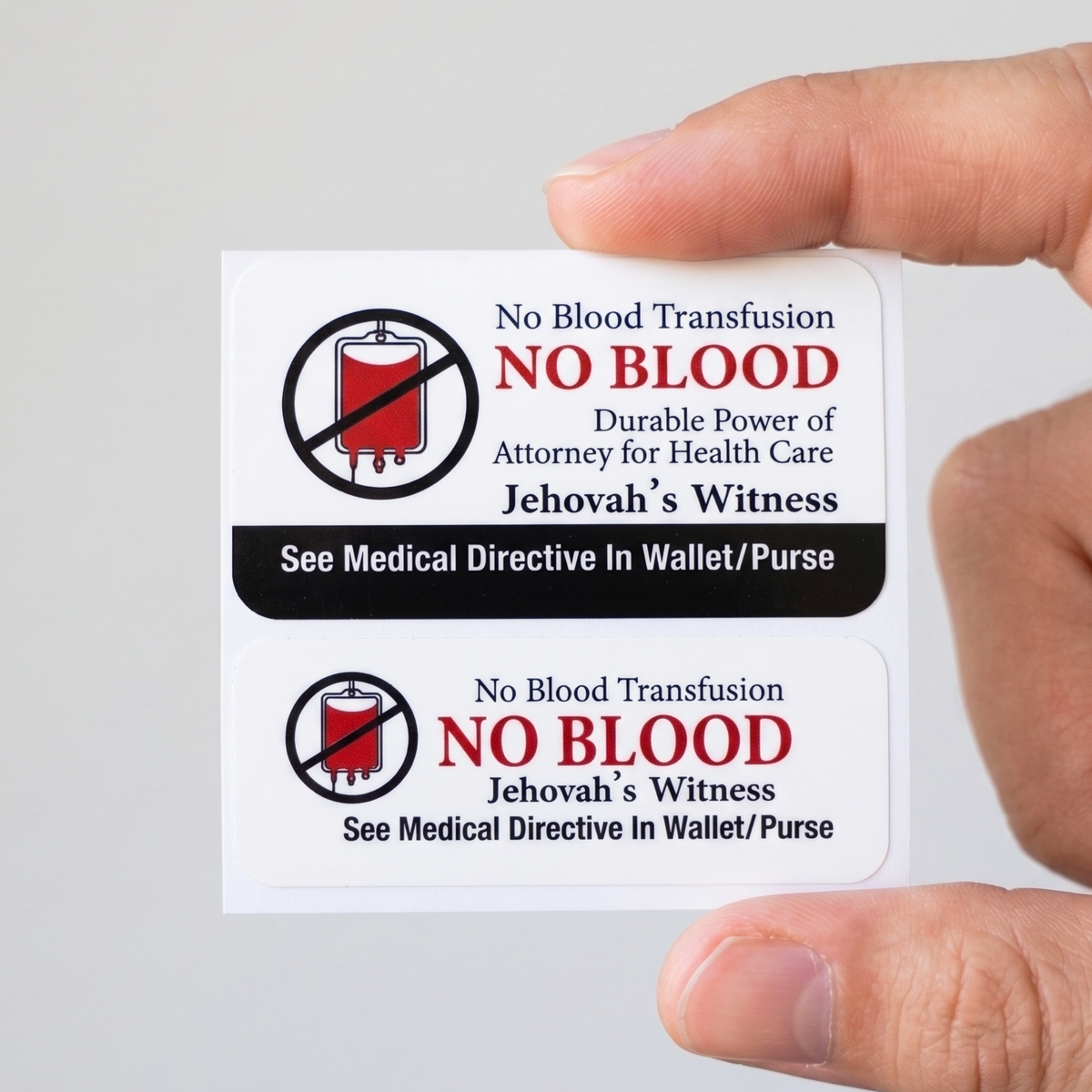 A hand holds two rectangular white stickers with printed religious/legal text and symbols reading 'NO BLOOD,' 'No Blood Transfusion,' and 'See Medical Directive In Wallet/Purse' on a plain light background.