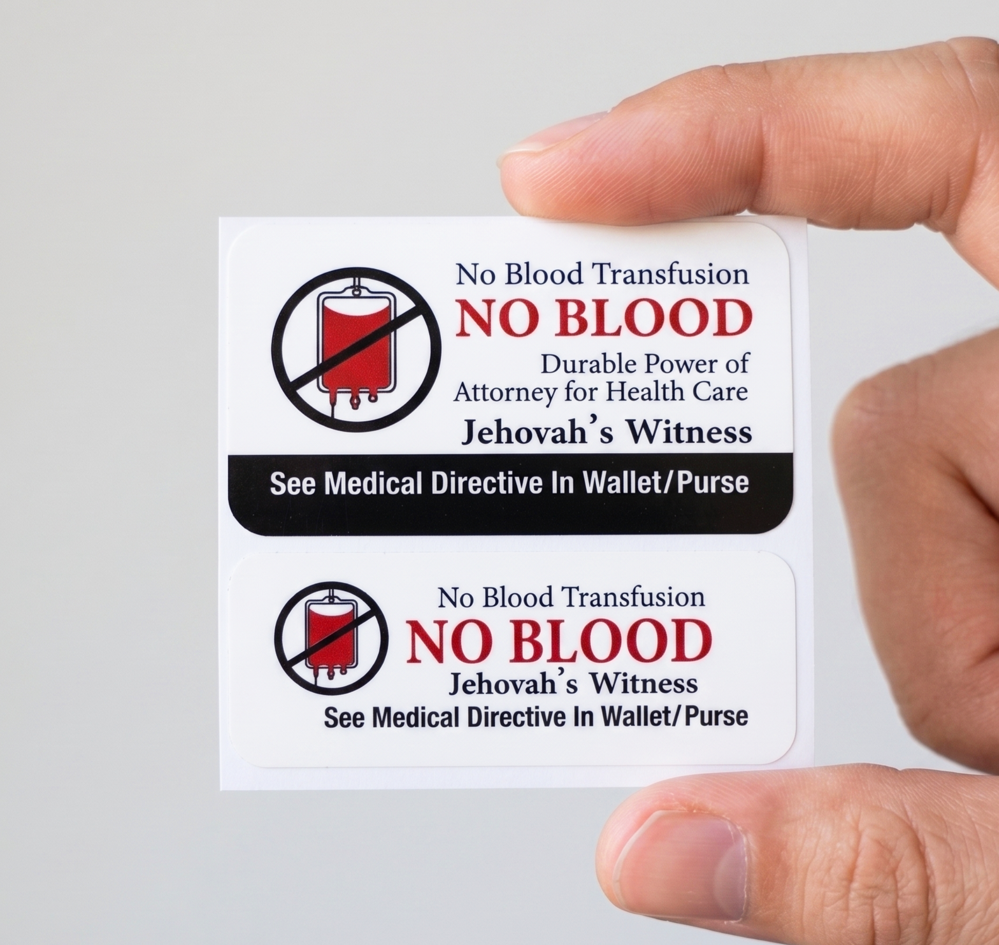 A hand holds two rectangular white stickers with printed religious/legal text and symbols reading 'NO BLOOD,' 'No Blood Transfusion,' and 'See Medical Directive In Wallet/Purse' on a plain light background.